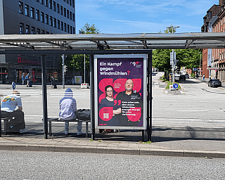 Plakat mit zwei Personen und Text über Windenergie an einer Bushaltestelle.