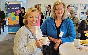 Zwei Frauen mit Tassen in einem Networking-Event, im Hintergrund weitere Teilnehmer.